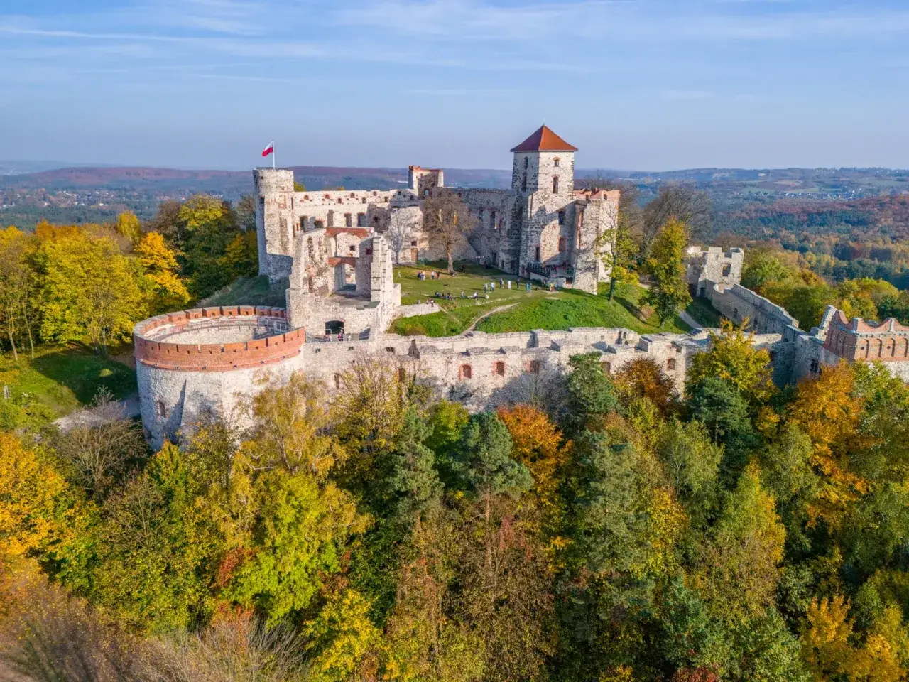 Ruiny zamku na Szlaku Orlich Gniazd otoczone jesiennymi drzewami. Biało-czerwona flaga powiewa na wieży.