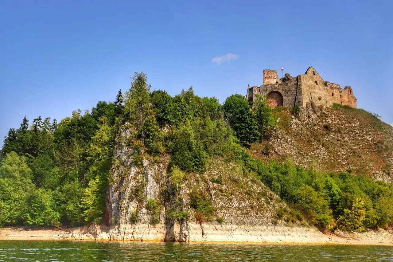 Zamek Czorsztyn góruje nad jeziorem, otoczony zielenią. Ruiny zamku na skalistym wzgórzu, z polską flagą.