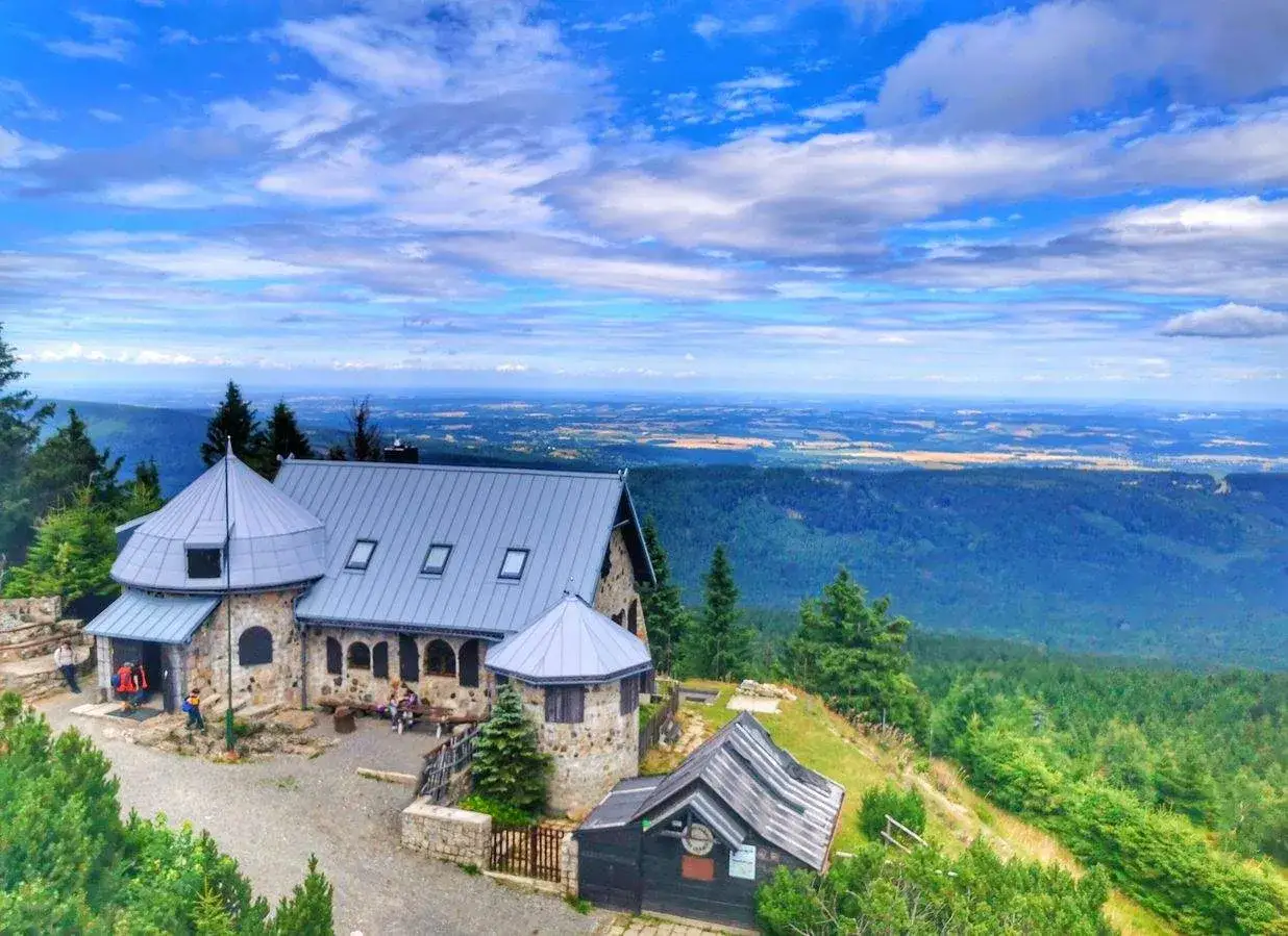 Kamienny budynek z szarym dachem na tle rozległej panoramy Gór Izerskich. Wokół zielone lasy i błękitne niebo z chmurami.