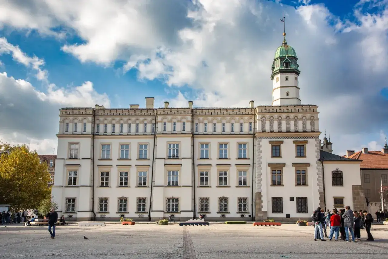 Rynek na Kazimierzu w Krakowie. Biały budynek z wieżą i zielonym dachem, plac z kilkoma osobami.