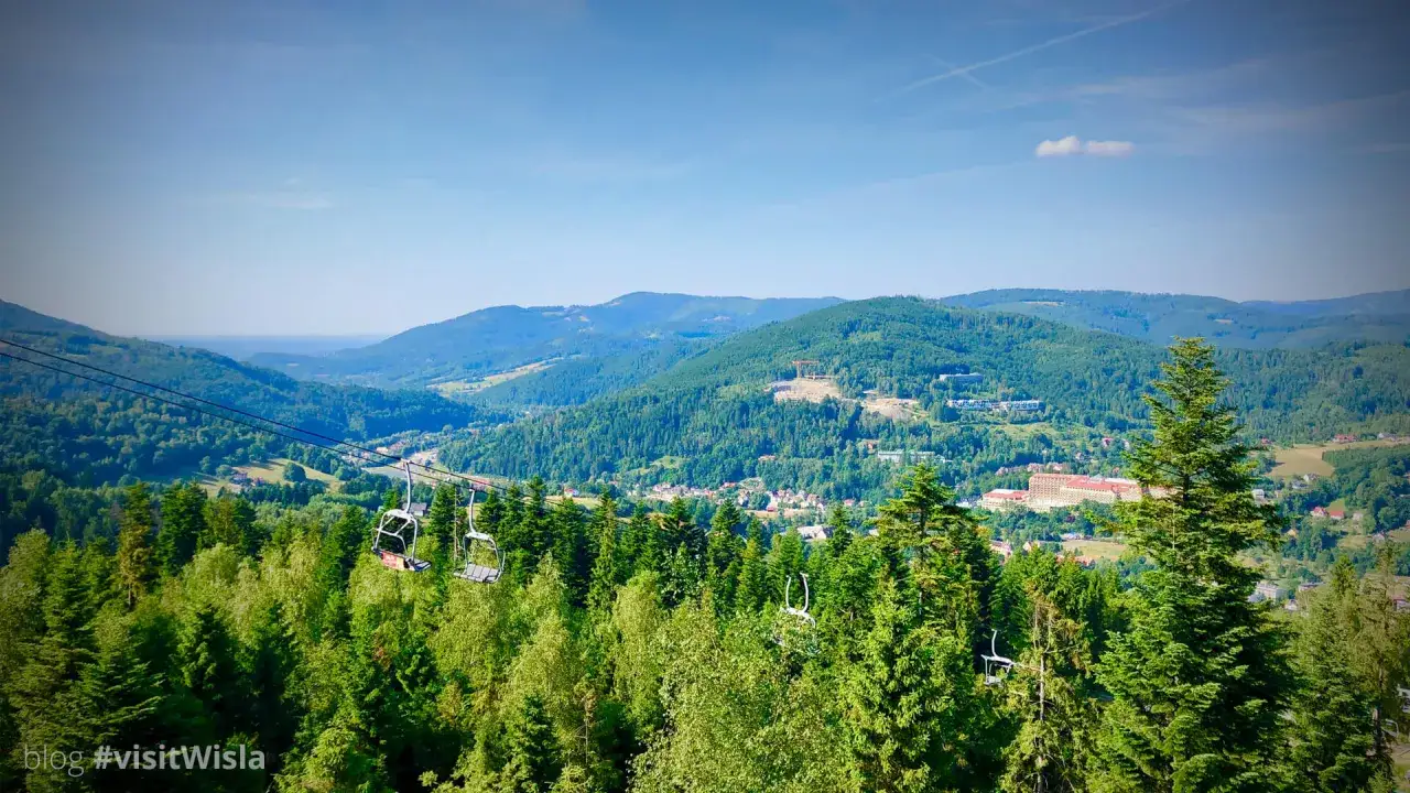 Widok na zielone góry w Wiśle, z widocznymi wyciągami krzesełkowymi i zabudowaniami w dolinie.