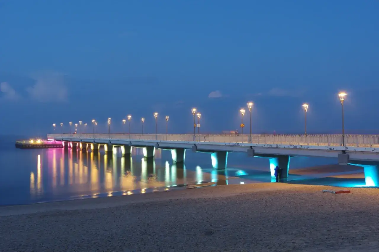 Molo w Kołobrzegu nocą, oświetlone latarniami i kolorowymi światłami, odbija się w spokojnej wodzie.