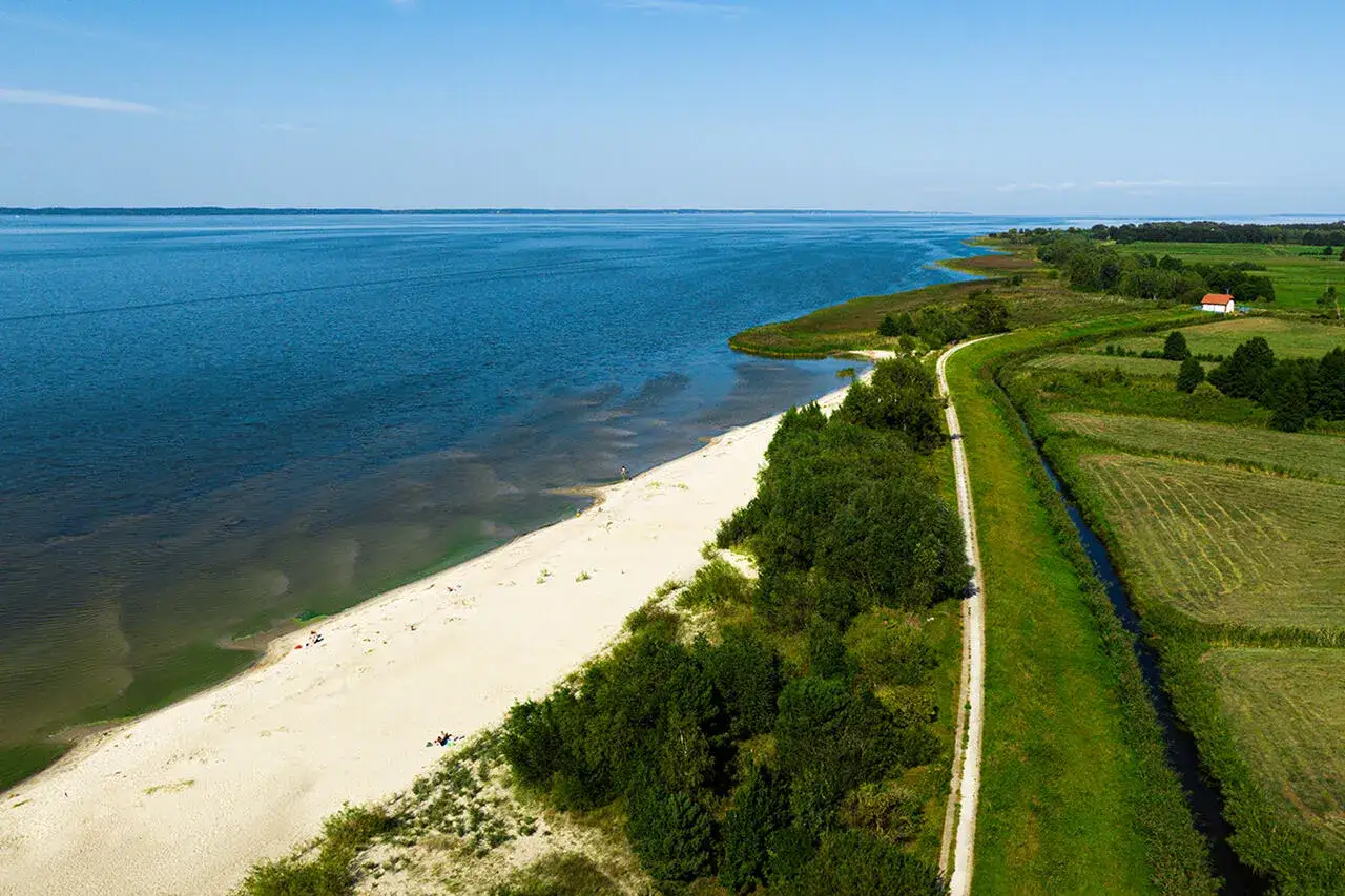 Piękny, piaszczysty brzeg nad Zalewem Wiślanym w Elblągu. W oddali widać pola i mały domek.