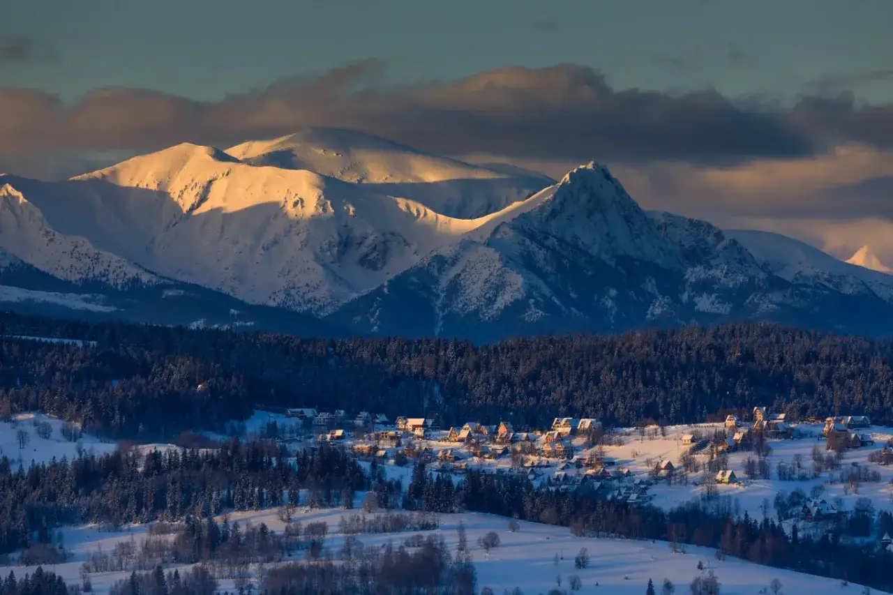 Zimowy krajobraz z ośnieżonymi szczytami Tatr i przysiółkiem Łapszanki. Słońce oświetla zbocza, tworząc złote refleksy.
