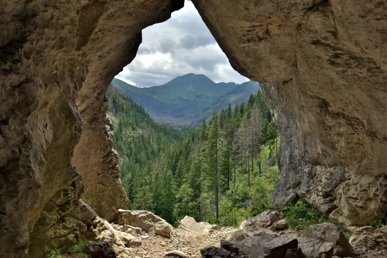 Widok z jaskini na Tatry. Skalny otwór, niczym okno, ukazuje zieloną dolinę pełną drzew i majestatyczne szczyty.