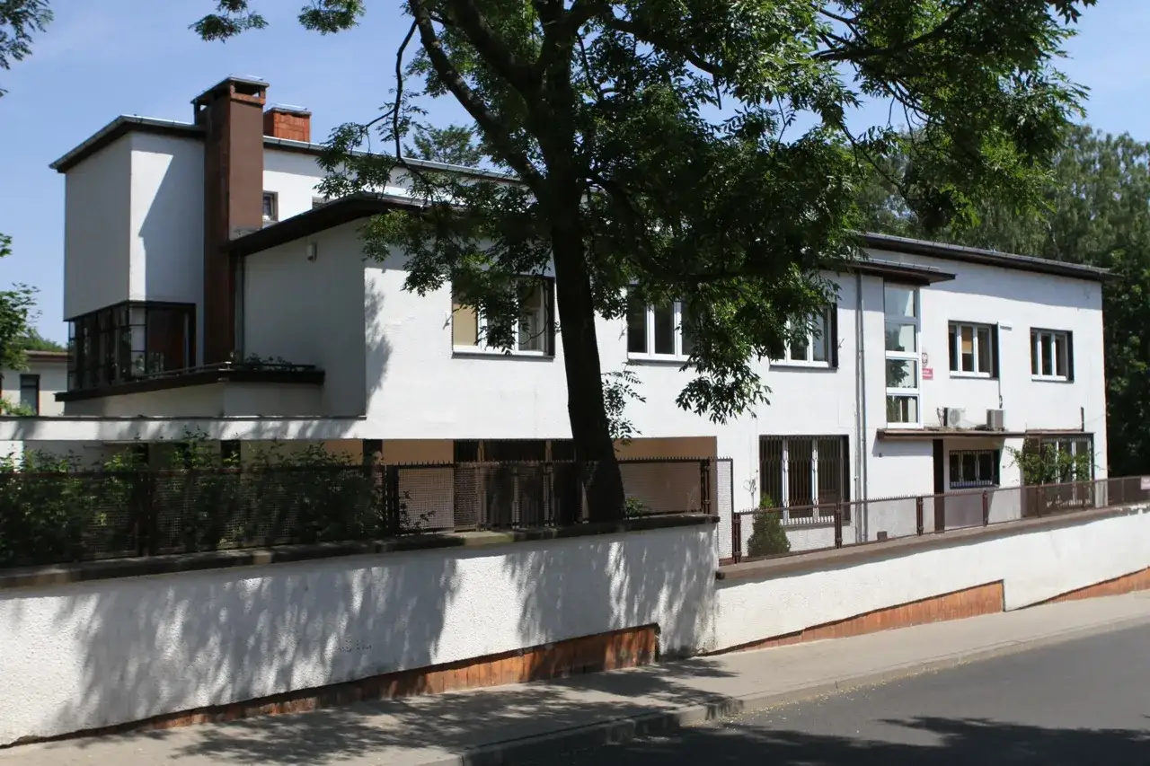Biały, modernistyczny budynek z ceglanym kominem, otoczony zielenią. To urokliwe miejsce w Gdyni Stare Miasto.