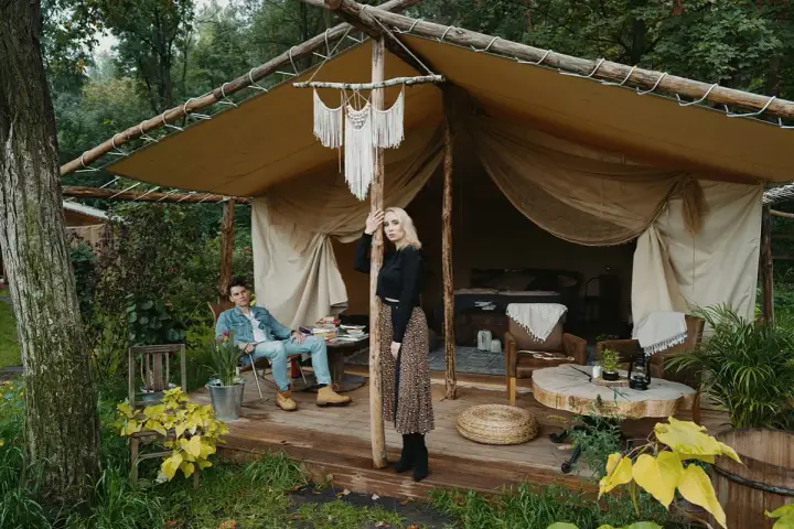 Para młoda relaksuje się w luksusowym kimura glamping namiocie. Mężczyzna siedzi na krześle, kobieta stoi przy drewnianym słupie.
