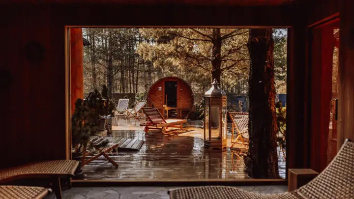 Drewniany domek w lesie, sauna beczka, leżaki i latarnia na mokrym tarasie.